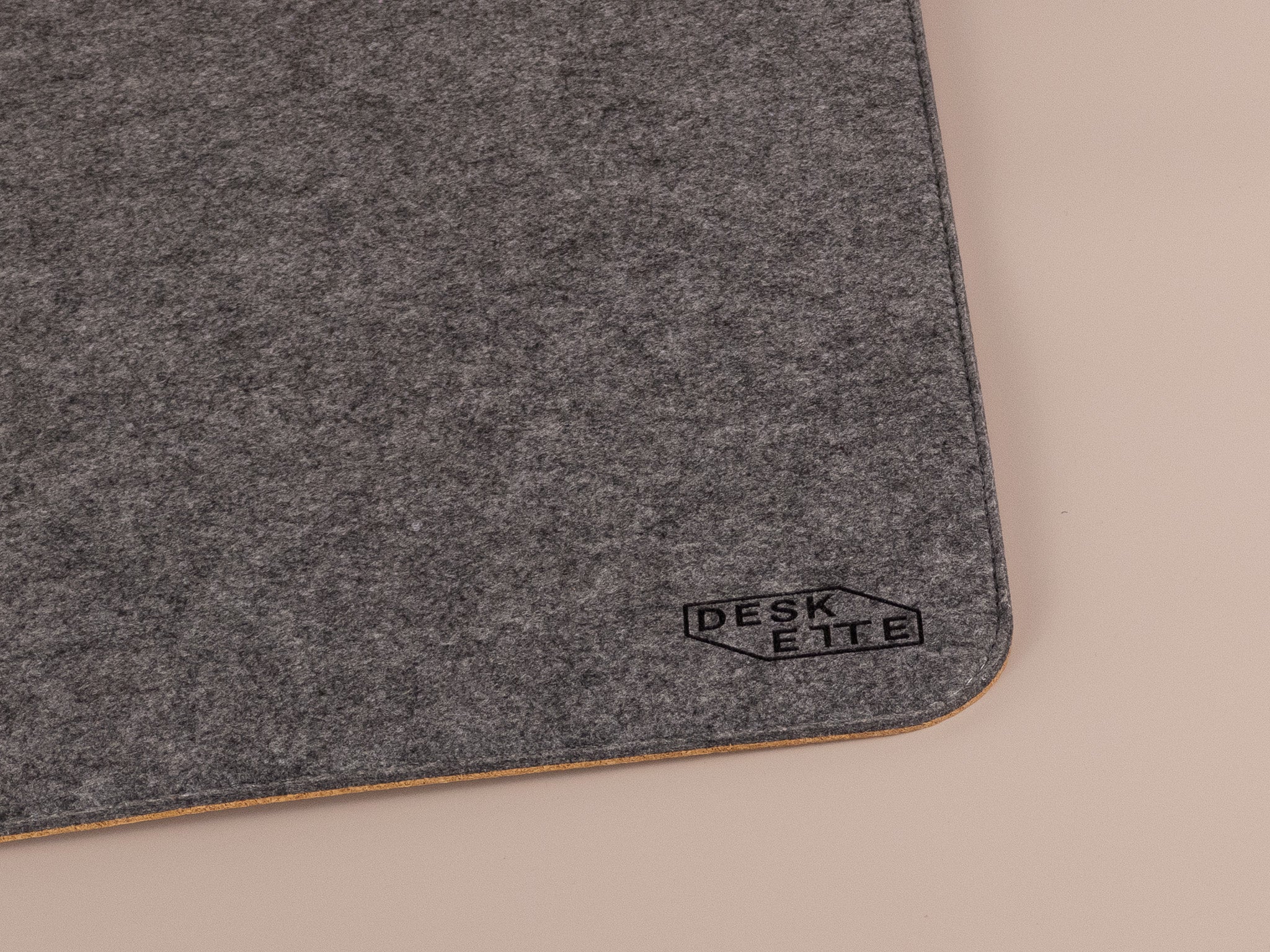 Close-up of Deskette logo on dark grey felt and cork desk mat corner, minimalist design