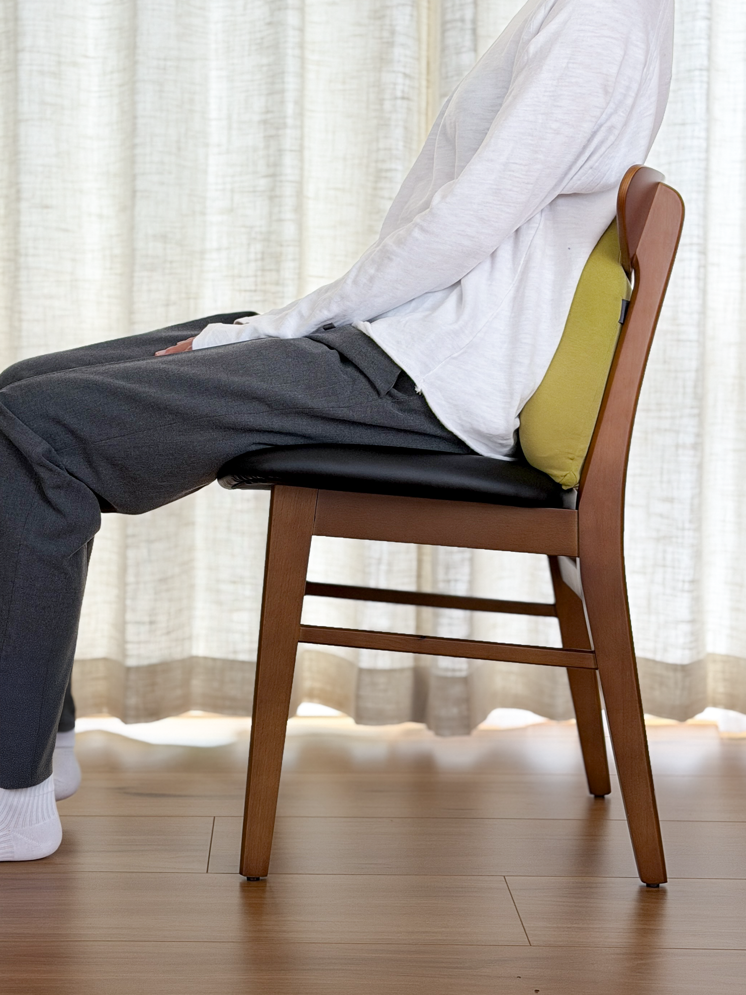 Person sitting on a wooden chair with a cushion, wearing a white shirt and dark pants, in a room with light curtains.