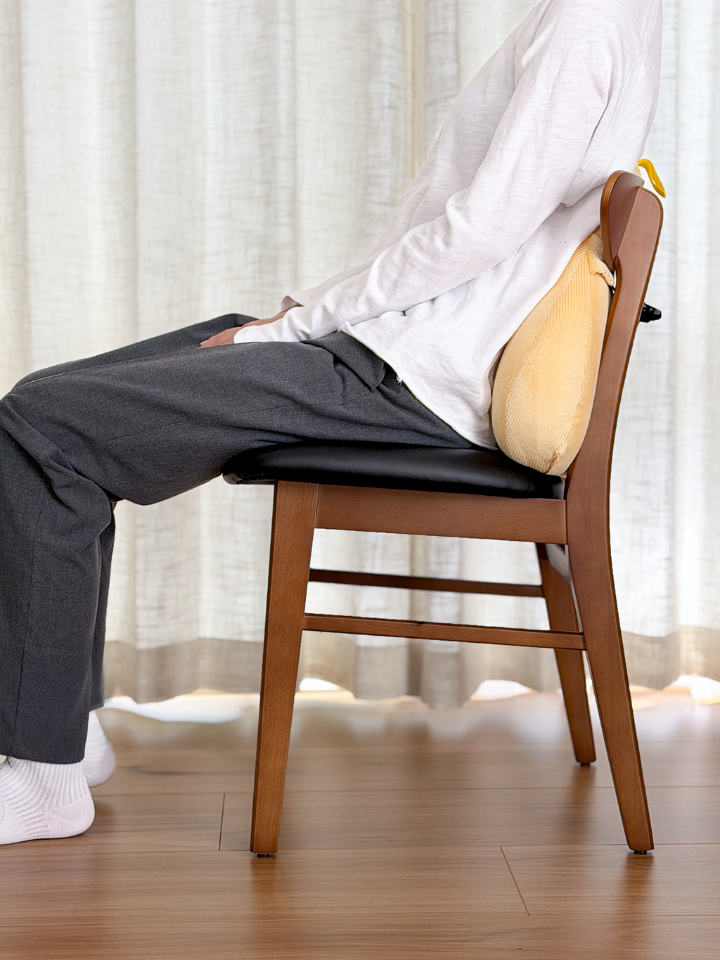 Person sitting on a wooden chair with a cushion, wearing a white shirt and dark pants, against a light curtain background.