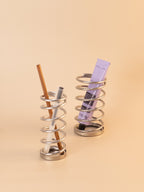 Metallic spiral holder with a pencil, pen, and tube of cream on a beige background